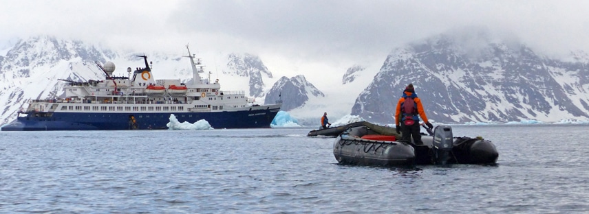 A guide drives an inflatable skiff through the water towards the ocean adventurer expedition ship floating among ice bergs in the Arctic.
