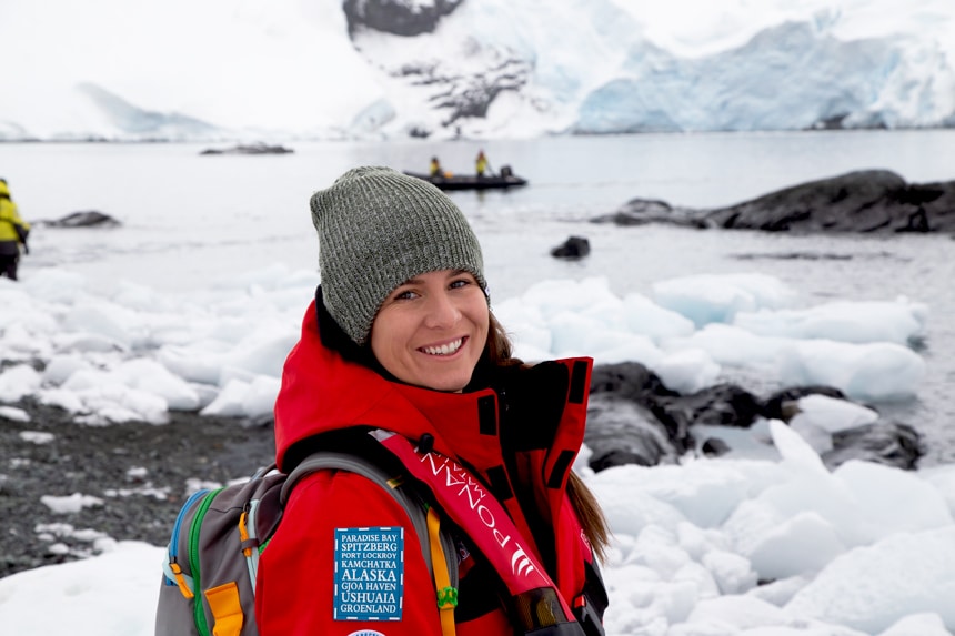 An Antarctica cruise passenger wearing a backpack and red parka poses for a portrait in front of an icy Antarctic landscape.