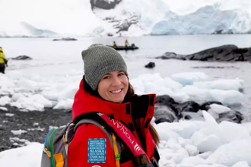An Antarctica cruise passenger wearing a backpack and red parka poses for a portrait in front of an icy Antarctic landscape.