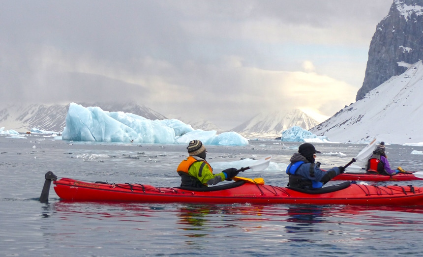 Arctic cruise guests in jackets and beanies paddle red double kayaks through the icy ice berg filled waters of Svalbard.