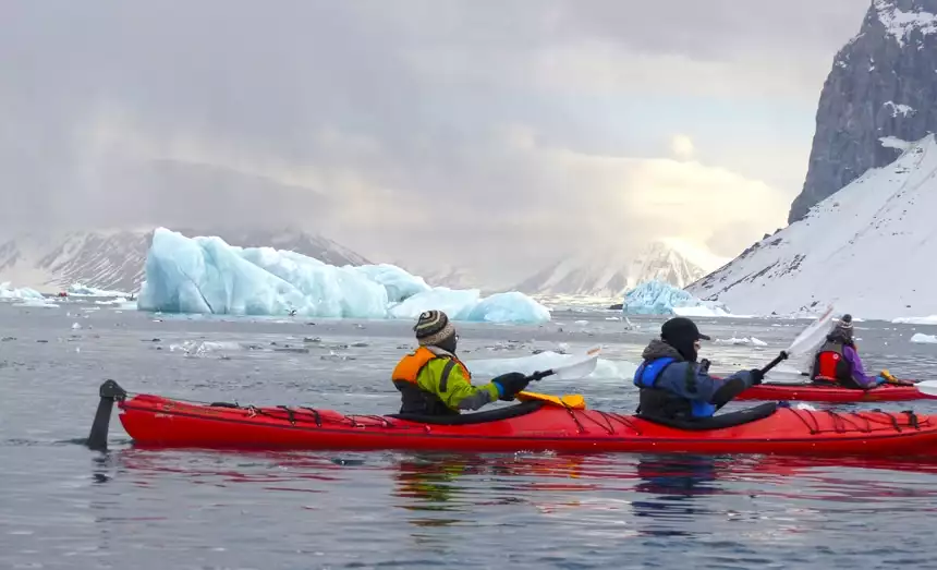 Arctic cruise guests in jackets and beanies paddle red double kayaks through the icy ice berg filled waters of Svalbard.