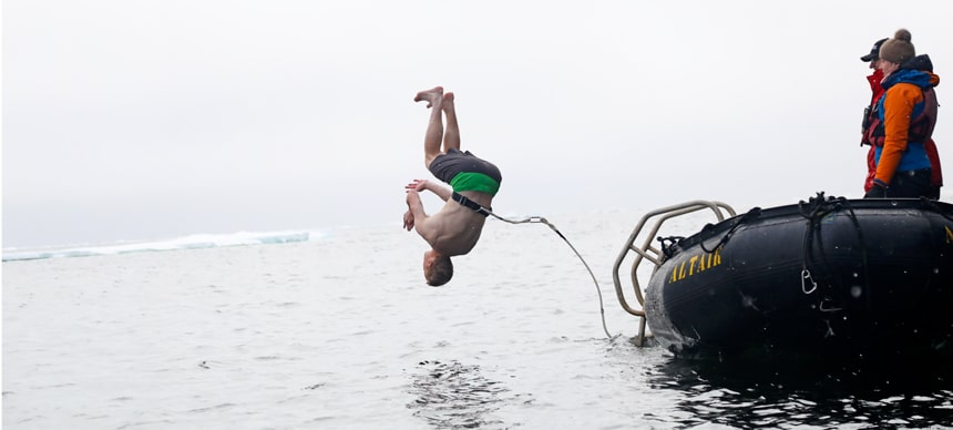 Polar plunge! A man flips into the arctic ocean in front of an ice shelf. He is harnessed to a black inflatable skiff for safety.
