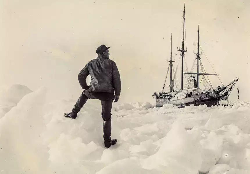 A vintage 1893 photo of famous explorer Nansen stands on shore with one foot on an ice berg staring at the Fram expedition ship stuck among thick icebergs.