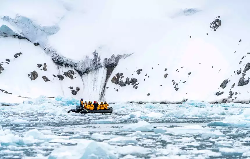 A black inflatable skiff filled with passengers in yellow parkas navigate the ice berg filled waters of the Arctic against a snowy shoreline.