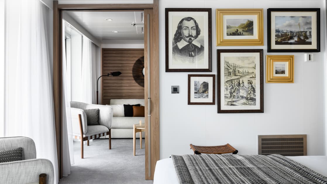 Owner's Suite aboard Le Dumont D'Urville luxury expedition ship, showing king bed, separate living room, bright white decor & photos & drawings of explorers on the walls.