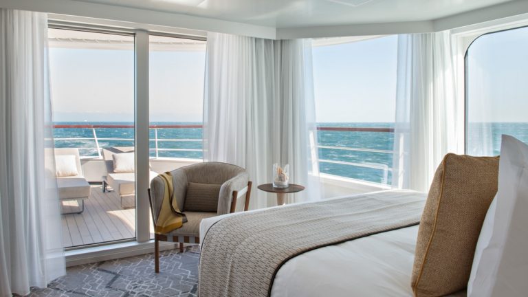 Floor-to-ceiling windows reveal the ocean & a terrace outside a white-decorated deluxe suite on Le Jacques Cartier French ship.