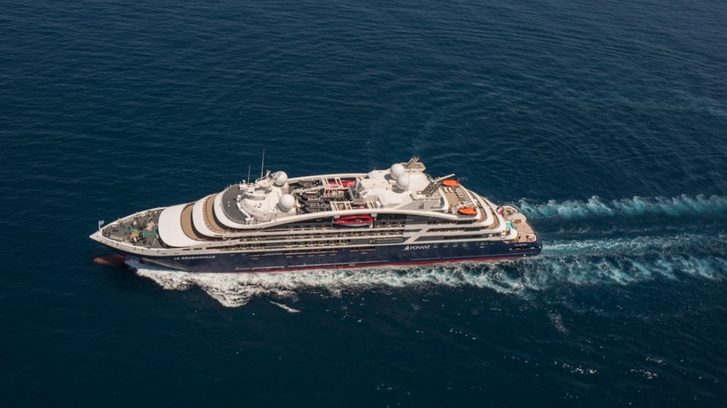 Aerial view of Le Jacques Cartier luxury expedition ship cruising in open waters, with dark blue & red hull & white upper decks.