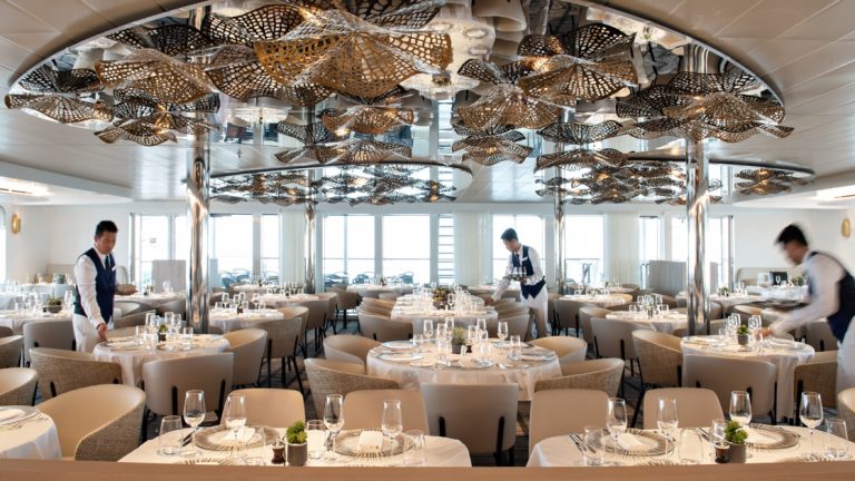 Dining room with elegant white tablecloths & floral gold light fixtures, being set for dinner by servers aboard Le Bellot Ponant ship.