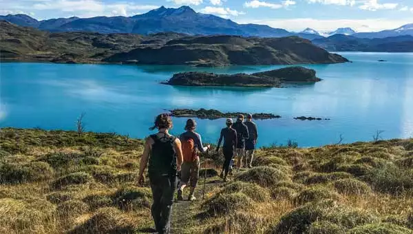 A line of hikers follow the trail through a grassy meadow down to a glacial lake with jagged peaks in the background, commonly seen on Chile Patagonia tours.