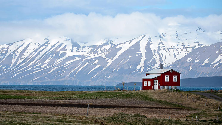 Small red house sits on stilts overlooking blue water with snowcapped peaks in the background, seen on an Iceland cruise.