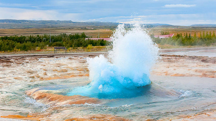 Geothermal pool explodes with icy-blue hot water amidst tangerine sand & sediment in Iceland.