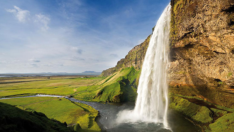 Large waterfall cascades into a small pool amidst bright green fields in Iceland.