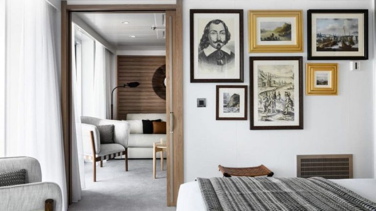 Owner's Suite aboard Le Bellot luxury expedition ship, showing king bed, separate living room, bright white decor & photos & drawings of explorers on the walls.