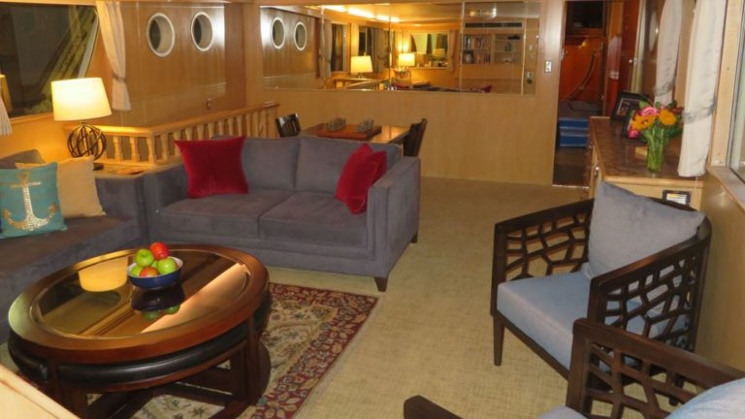 Main salon aboard the Sea Star yacht, with short blue couches, padded blue chairs, wooden coffee table, large view windows & wooden motif.