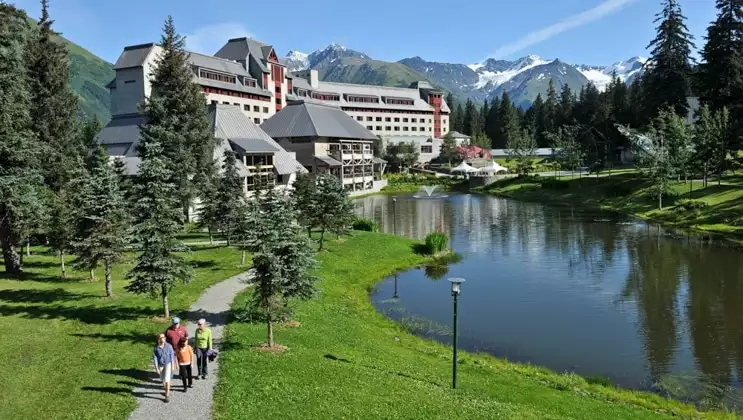 Aerial view of Hotel Alyeska, a chateau-style, multi-story gray hotel set among lush grass, a pond & mountains, on a sunny day.
