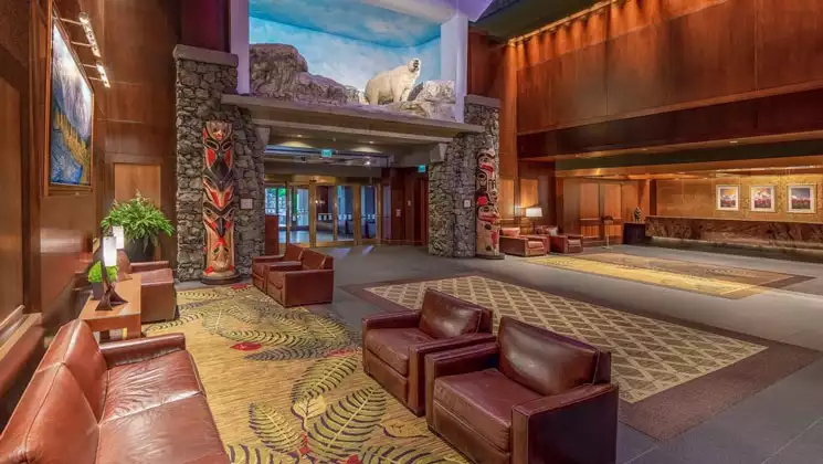 Lobby of Hotel Alyeska, with leather chairs & sofas, wooden walls, totem poles & stuffed bear, plus soft lighting & large entryway.