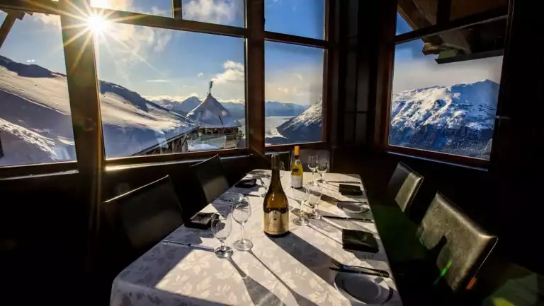 Table set for 4 with white tablecloth, classy dinnerware & panoramic view overlooking snowy mountains & lake below, at Hotel Alyeska.