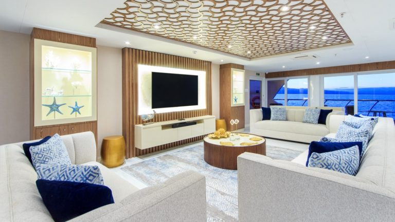Living room on mc Elite Galapagos ship, with white couches circling a flatscreen TV & small round coffee table, with view windows behind.