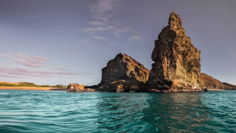 Turquoise water beside white-sand beach & 2 large rock outcrops reaching to the sky, seen on the Elite Galapagos cruise.