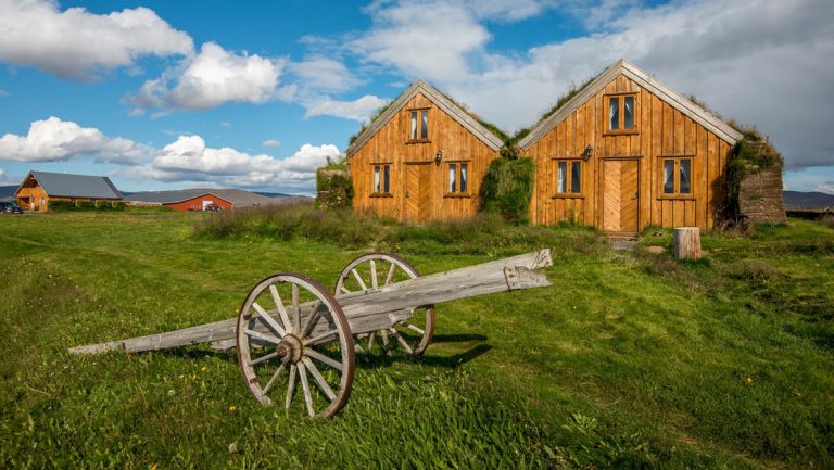 Wooden guesthouses with sod roofs in a grassy field with old farm equipment in front, on a sunny day at Fjalladyrd Guesthouse in Iceland.