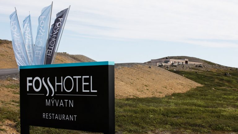 Outdoor hotel sign in dark brown with flags rising behind, advertises the Fosshotel Myvatn, with building in background & green hills in front.