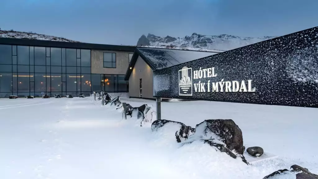 Exterior ground-level view of Iceland Hotel Vik i Myrdal in winter at dusk, with glass walls & illuminated black rectangular sign.