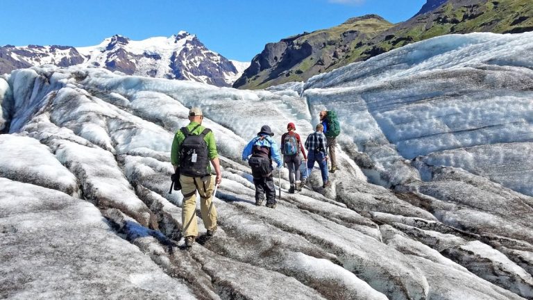 Travelers trek atop a dirty white & blue glacier on a sunny day during the Iceland Adventure: Land of Fire & Ice tour.