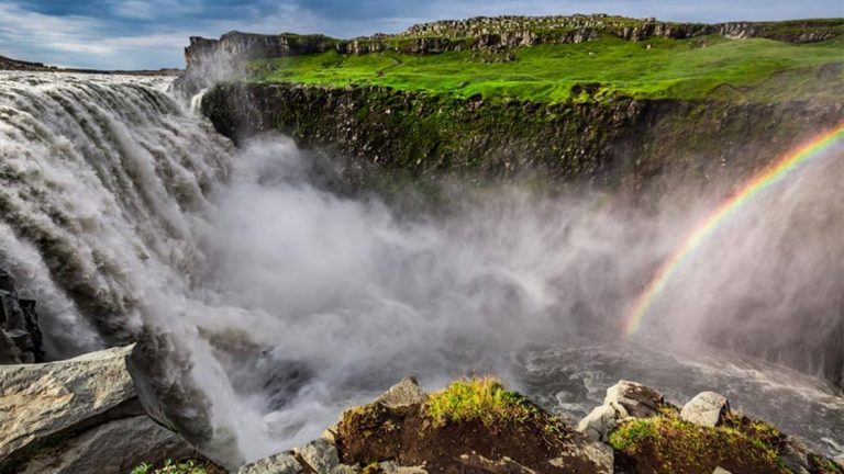 Huge waterfall sends rushing water down past lush green hillsides, with spray from the water creating a rainbow on the Iceland Adventure.