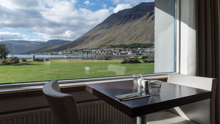 View out window from 2-top table with padded chairs in Iceland Hotel Isafjordur restaurant, looking at fjord & green grass.
