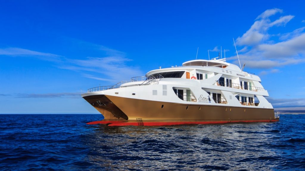 Exterior of mc Elite Galapagos catamaran with gold hull & 3 white decks, cruising in calm open water on a sunny day.