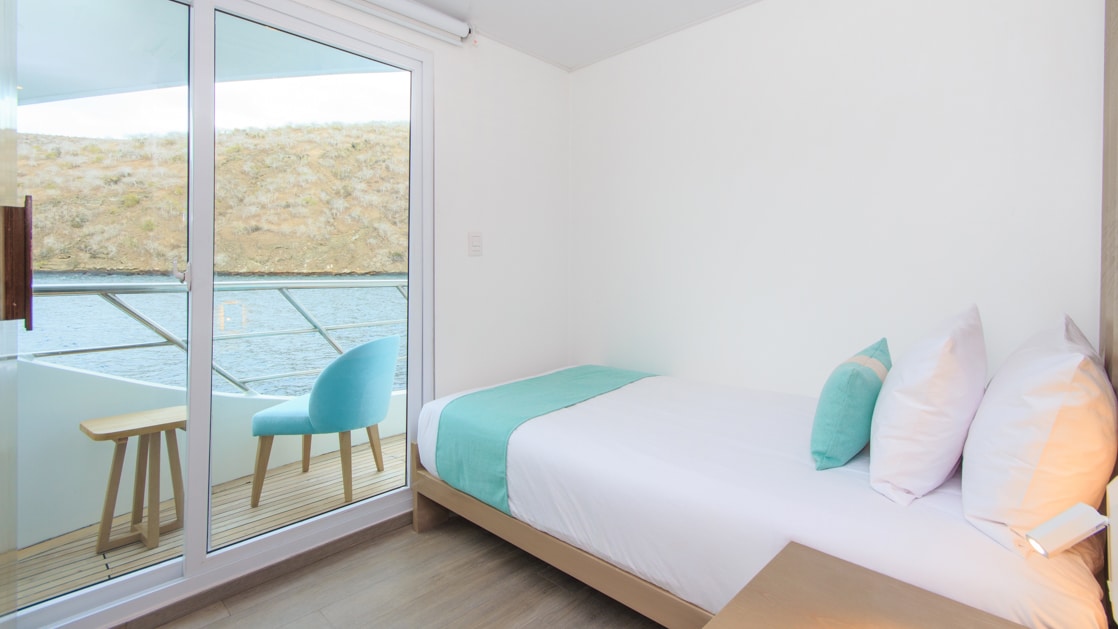 Twin bed with white, beige & teal bedding in slender room with white & wood accents & view onto glass sliding door & private balcony.