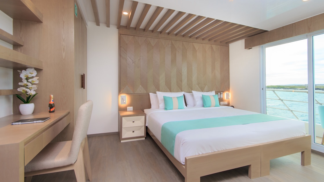 Double bed with white, beige & teal bedding in room with white & wood accents, wooden desk & shelves & glass door onto private balcony.