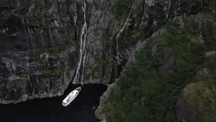 Aerial view of Sea Star small white yacht idling beside tall rock walls with waterfalls during a Kenai Fjords NP cruise.