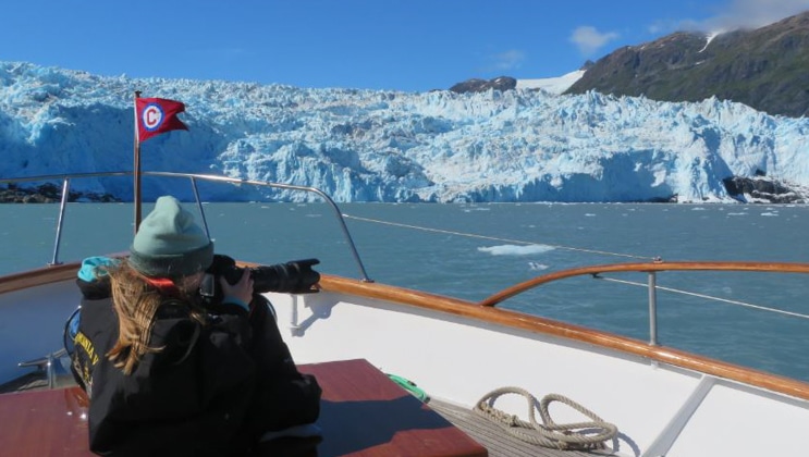Alaska traveler sits near bow of a small yacht with camera out, photographing an icy blue glacier on a sunny, clear day.