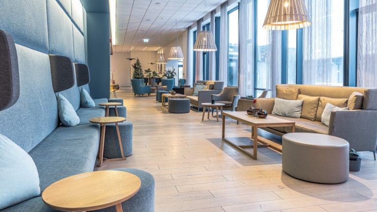 Lobby of Hotel Vik i Myrdal, with wall of teal couch, wooden coffee tables, wood flooring, gray couches, hanging lamps & floor-to-ceiling windows.