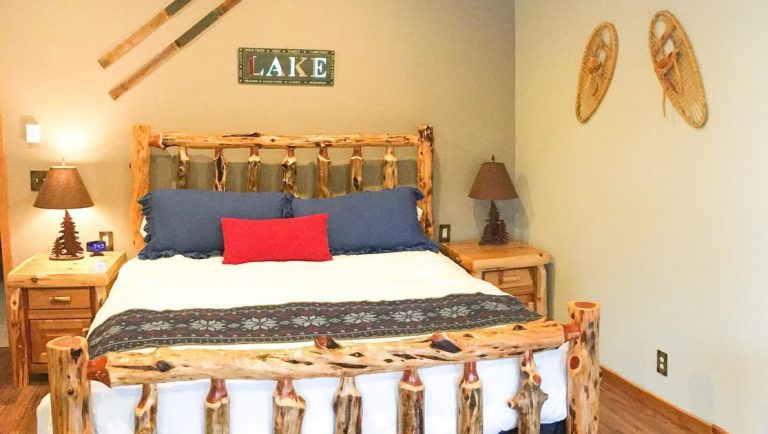 Modern & rustic log bed covered in quality linens with bedside tables & lamps in a modern cabin with snowshoes & old skis on the wall.