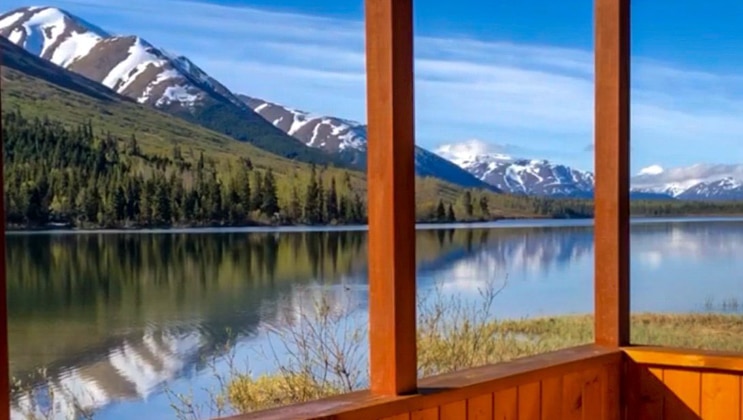 View from a lakefront cabin wooden porch at the Summit Lake Lodge, on a sunny day with glassy water reflecting the hills & mountains.