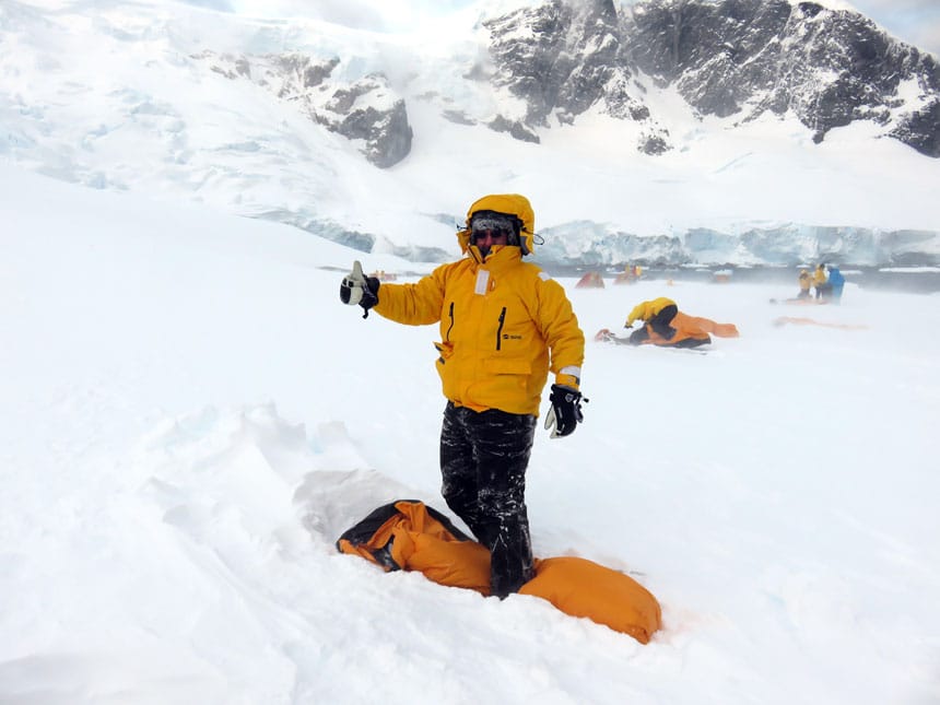 polar traveler in yellow jacket gives the thumbs up while standing over a yellow bivvy sack atop a snowfield while camping in antarctica.