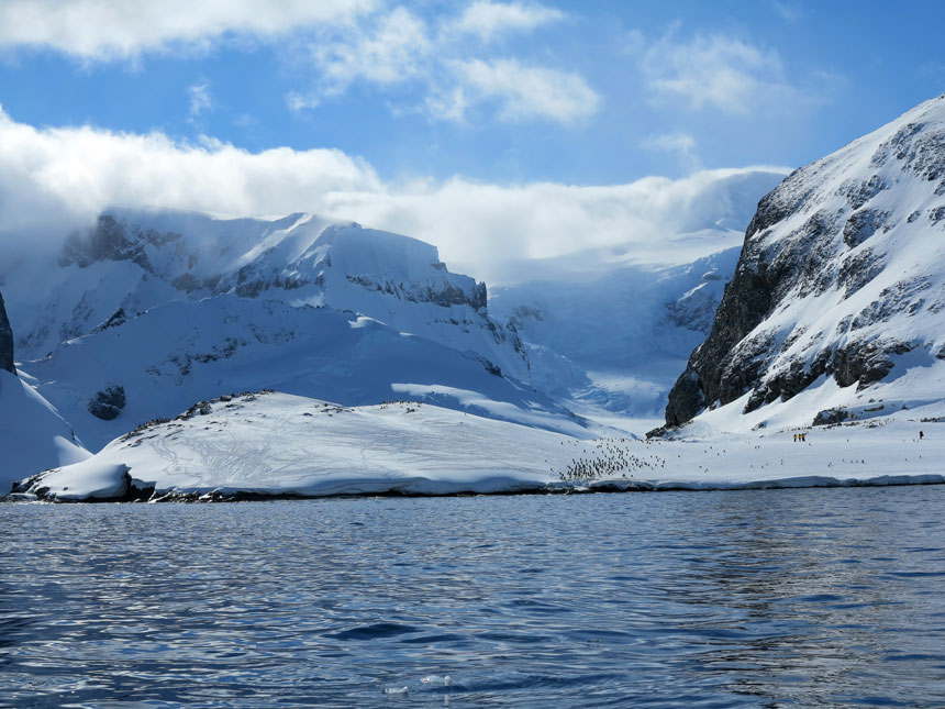 Cuverville Island covered in snow with dark cliff features & tiny specks of penguins on a sunny day with water in the foreground.