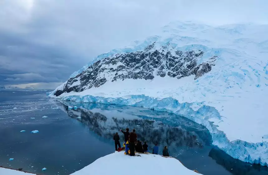 Neko Harbor, one of the places in Antarctica, with people perched atop a lookout point viewing the glassy water below on a cloudy day.
