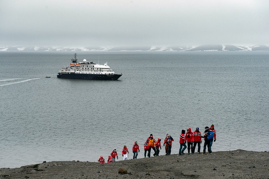 Guests wearing red parkas hike up a barren hillside in Antarctica as a blue and white ship floats in the dark ocean behind them.