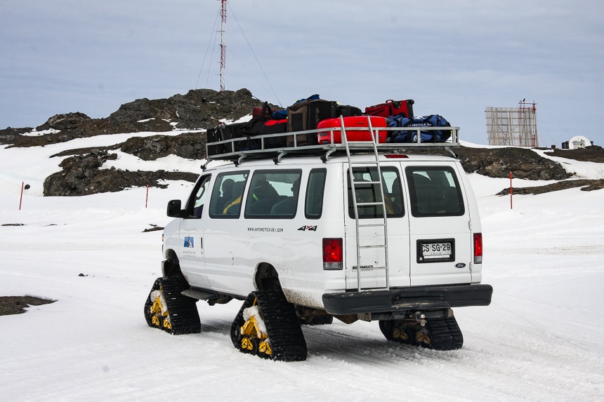 A white van with tracked triangle shaped tires for snowy Antarctic driving carries guests and their luggage from the ship to the air plane for an Air Cruise itinerary