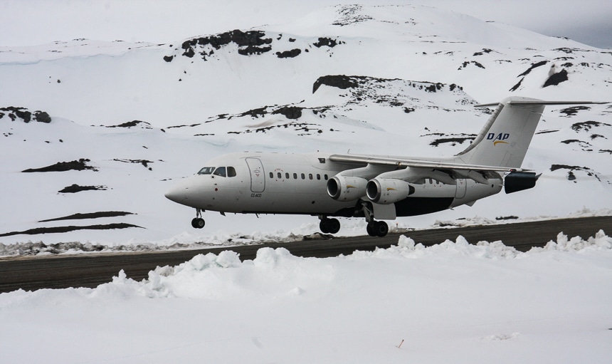 As part of an Air Cruise itinerary a white air plane lands on a grey run way in the middle of a snowy mountainous Antarctica landscape.