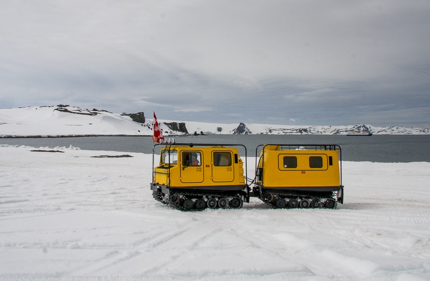 Against a grey Antarctica ocean landscape two yellow tank-like land rovers drive across the snow as part of an Air Cruise.
