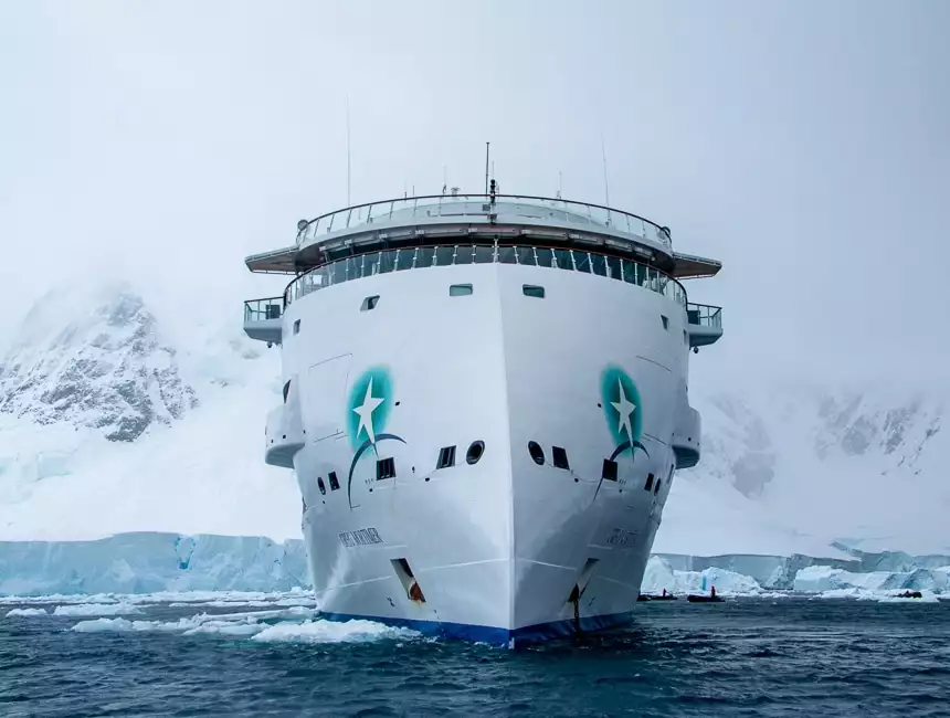 An all white futuristic and modern ship cruises the dark ocean water in front of a mist covered snowy mountain range in Antarctica.