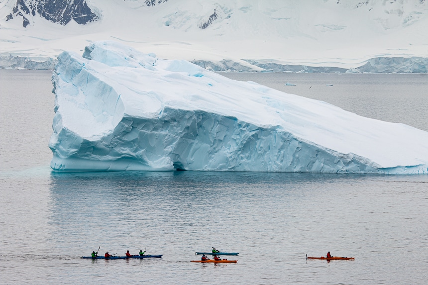 A group of kayakers are dwarfed in size as they paddle around a massive teal and white floating iceberg in Antarctica.