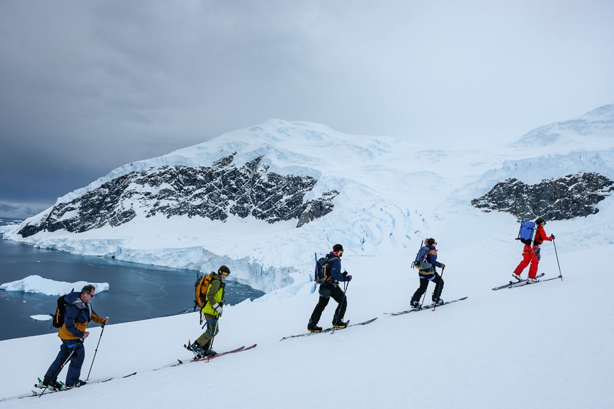 On a grey overcast day in Antarctica a group of guests ascend a snowy hillside on skis as part of an added cost activity provided by an active Antarctica cruise line.