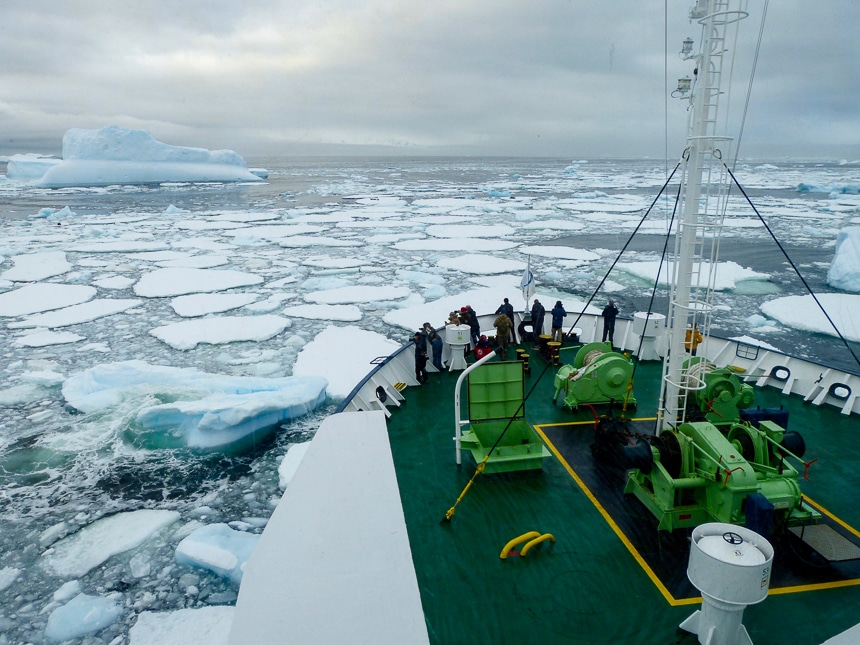 Guests gather at the bow of a ship as it breaks through a field of icebergs floating in the ocean of Antarctica.