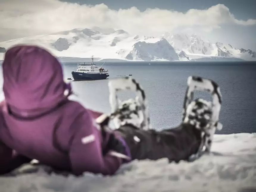 A guest wearing snowshoes lays on the snow, in the distance a blue and white ship navigates the polar ocean in front of white Antarctica mountain landscape