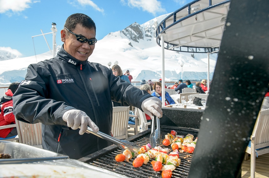 A happy crew member holds tongs and bbq's kebabs outside on a grill for a top deck outdoor dining experience in Antarctica.